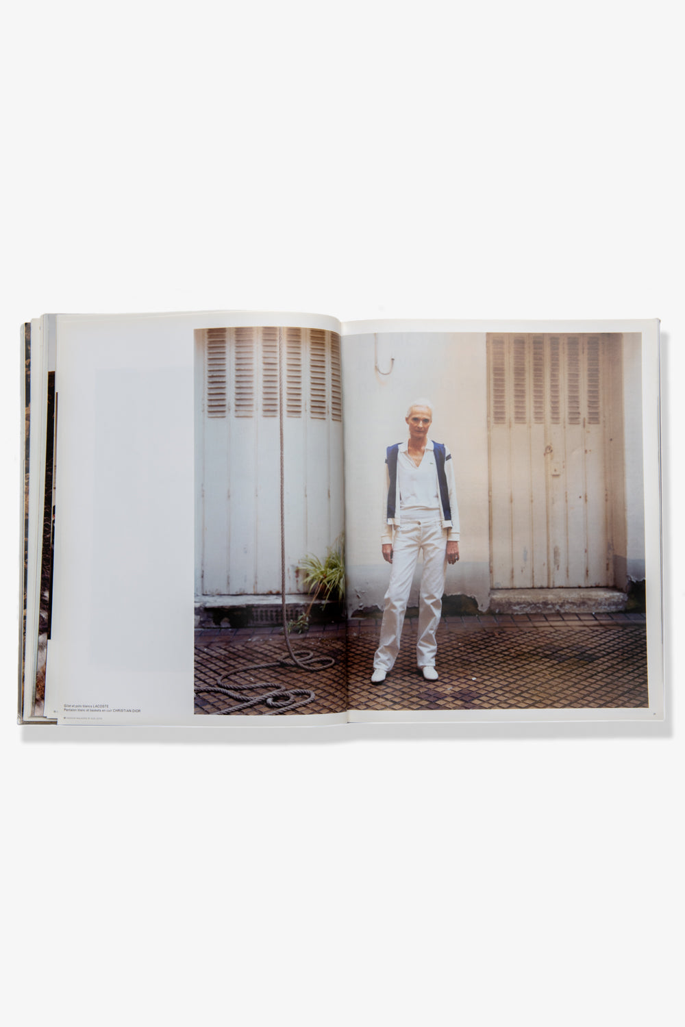 A photograph of a spread of Alec Soth's Paris, Minnesota. Recto: A male model stands against the exterior of a worn white building, in white pants and top, with a blue-sleeved sweater draped over his shoulders. Verso: A panel of the building. 