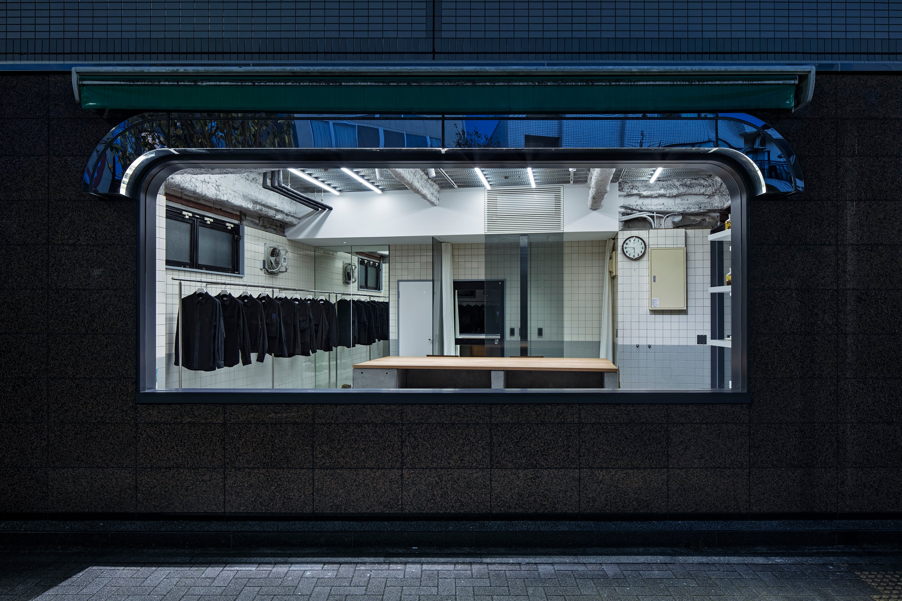 View of store from the outside into the store's large window. Store features long rail on left, a large wood table in the center and stark white overhead lights.