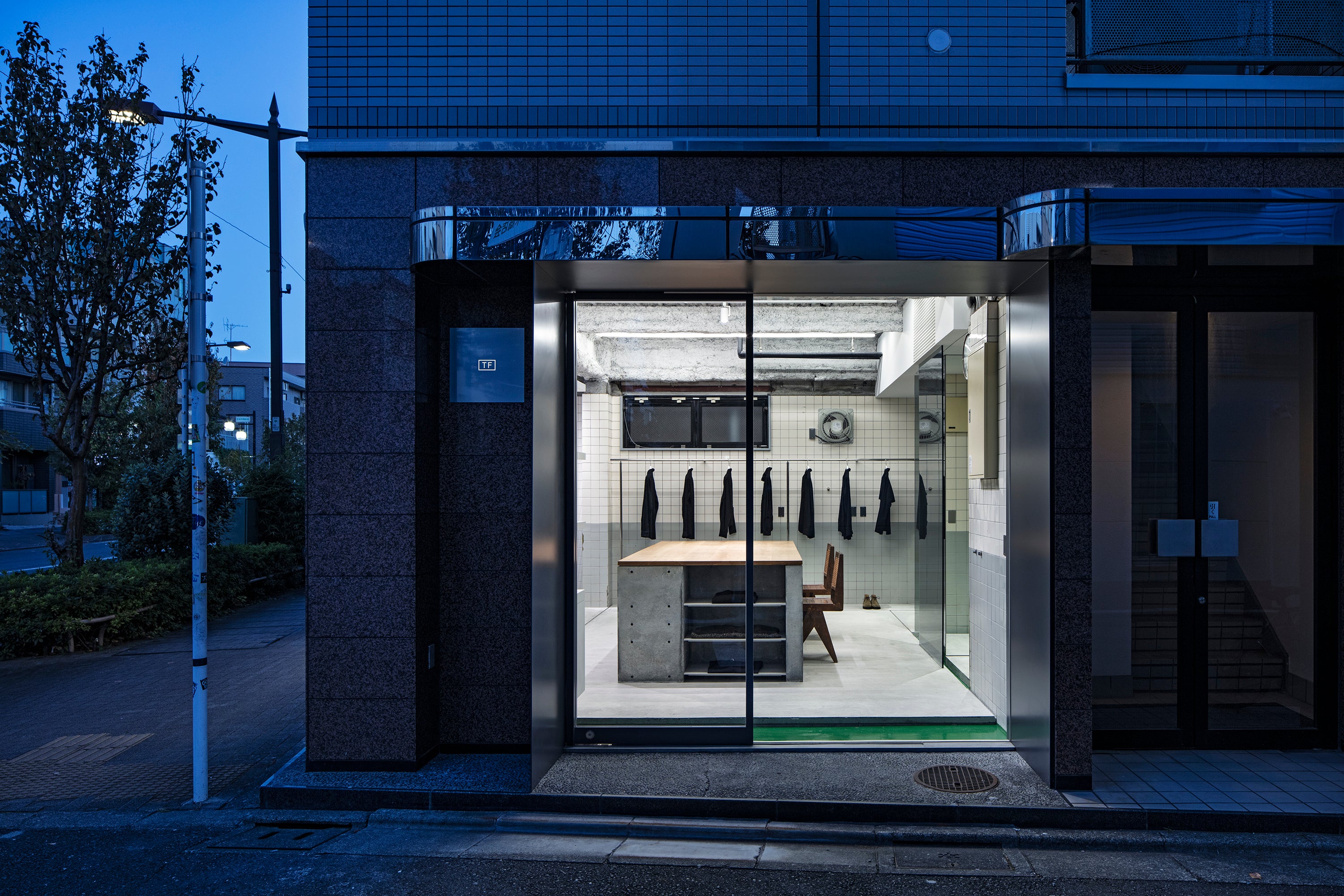 View of store from outside at dusk. The exterior is dark and inside is lit with bright overhead lights. Store shows large cement and wood top table with a clothing rail along the back wall.
