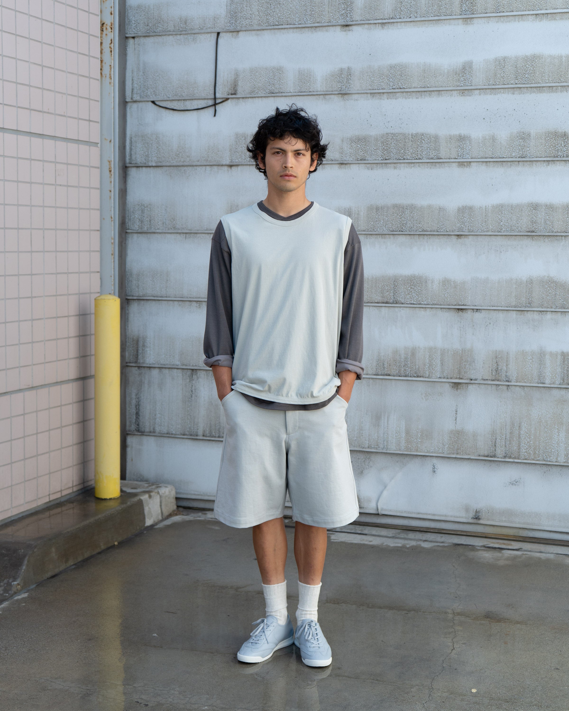Model wears a LW106 Sleeveless T-shirt in Swiss natural over a LW135 Football T-shirt in Grey Olive, and LW1150 Jersey Shorts in Swiss Natural and LW1070 German Military Trainer against a corrugated metal background