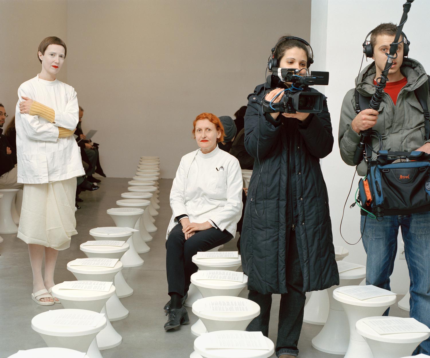 A still from Alec Soth's photography book Fashion Magazine: Paris Minnesota. From left to right, a woman stands in a white smock; an older woman in a white smock sitting; a cameraperson in a padded 3/4-length coat; her boom mic operator with gear draped around his front. 
