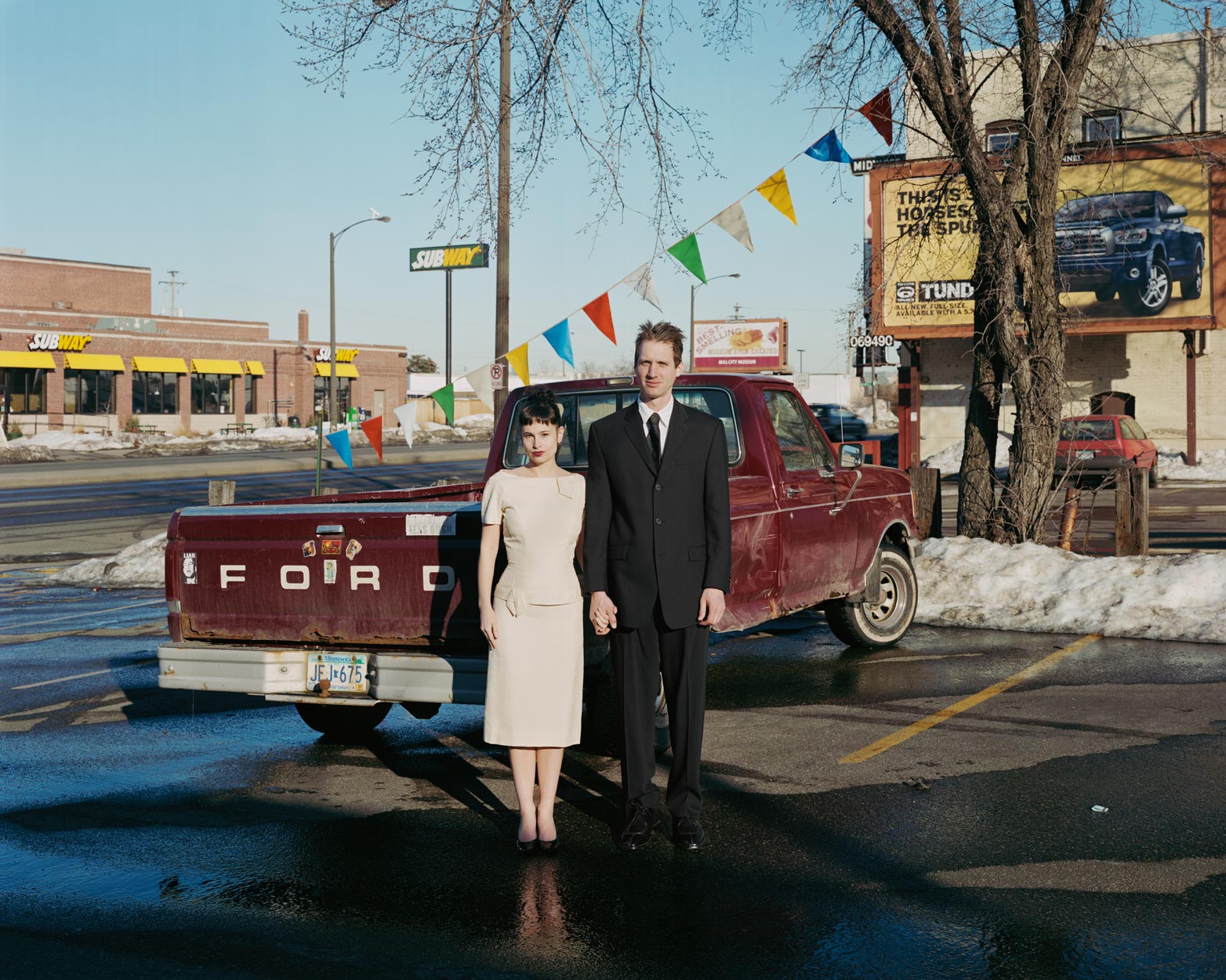 Photograph from Alec Soth's photography book "Paris Minnesota." A couple in formal attire stands in front of an old red Ford truck in a parking lot, a Toyota Tundra ad in the background.