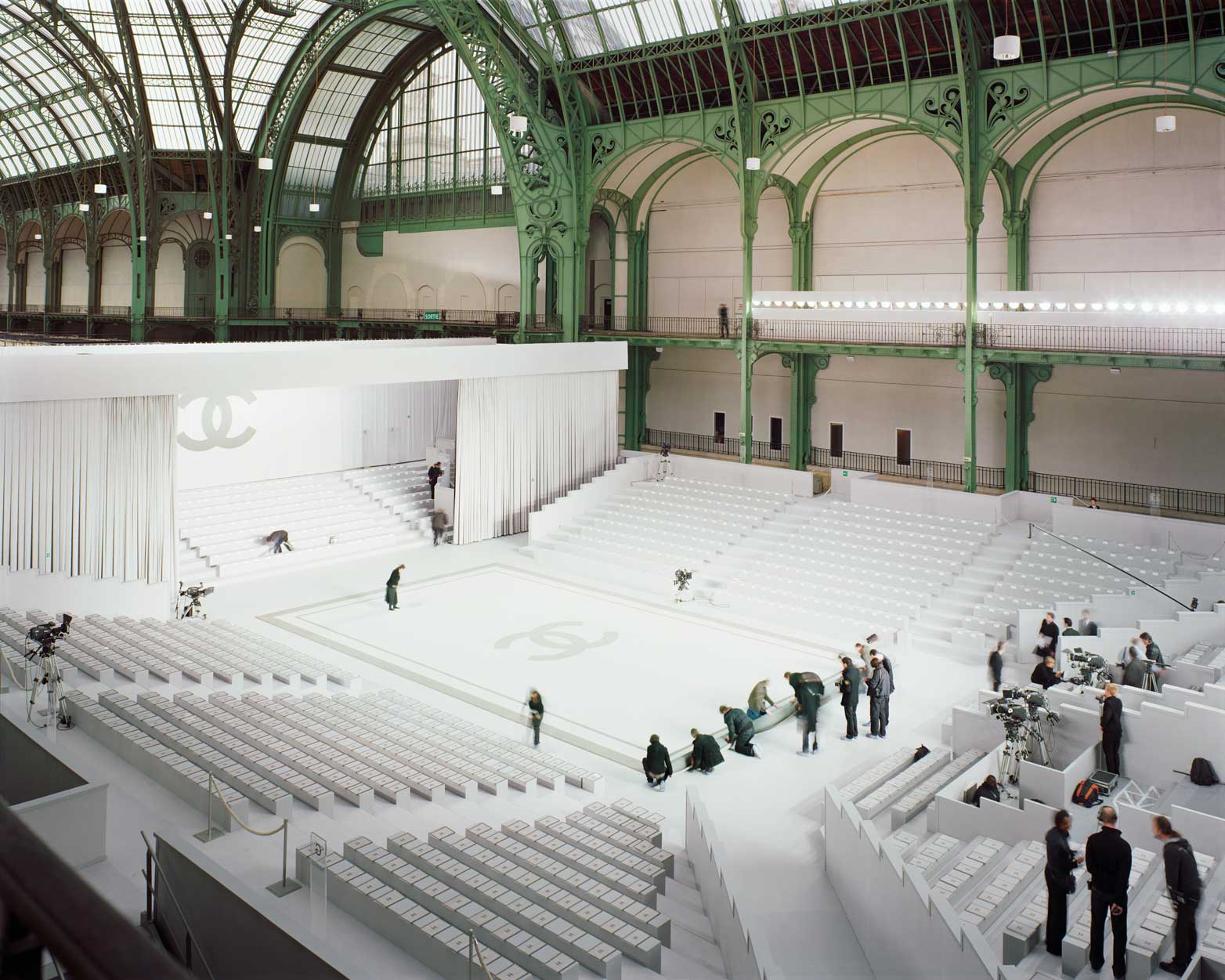 A still from Alec Soth's photography book Fashion Magazine: Paris Minnesota. The grand runway, the setting for a Chanel show, stands empty but for producers and stage-hands preparing for the runway. The staggered, theater-like seating sit empty. 