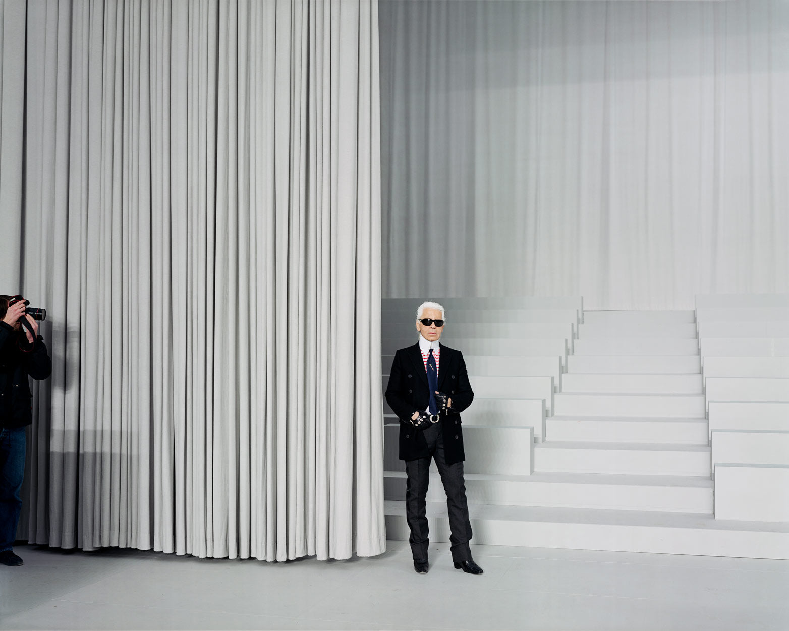 A still from Alec Soth's photography book Fashion Magazine: Paris Minnesota. The fashion designer Karl Lagerfeld, dressed in a black suit and wearing black sunglasses, sits at the edge of a drawn-back floor length grey curtain, with a short series of staggered bench seating behind him. Extending out of the frame, a photographer aims their camera at Lagerfeld.