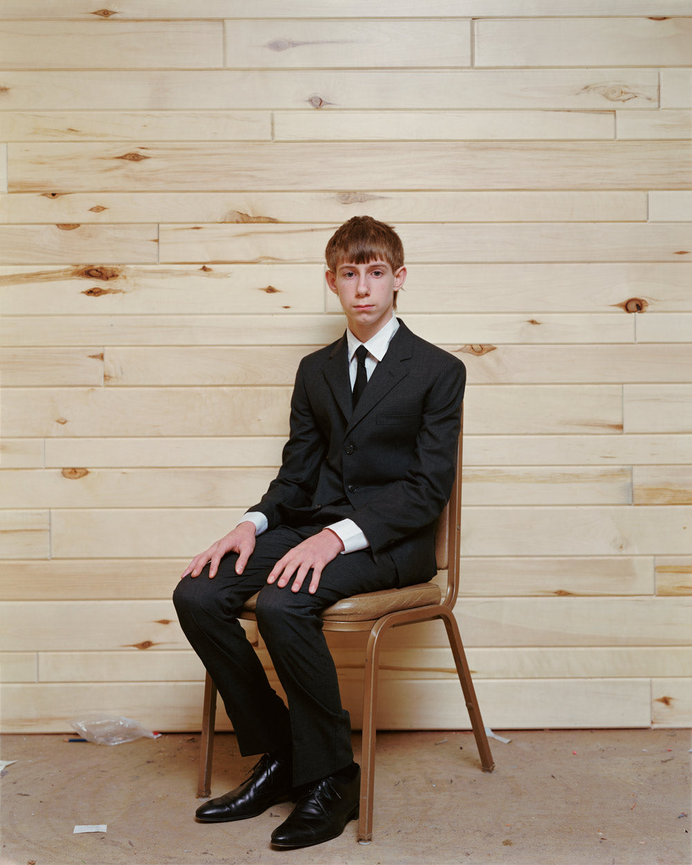 A still from Alec Soth's photography book Fashion Magazine: Paris Minnesota. A young man in a full suit sits on a plain brown side chair, askew from the direct sight line. He is seated against a wall made of pine or timber. His expression is blank and nonplussed.