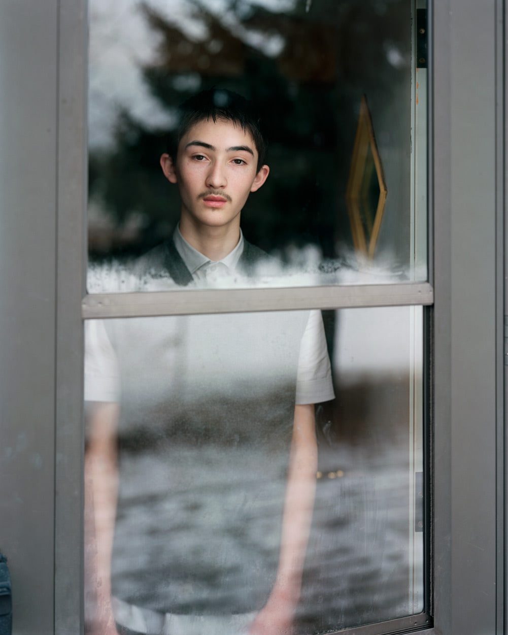 A still from Alec Soth's photography book Fashion Magazine: Paris Minnesota. A young man looks out of the window, his view reflected against the mirror behind which he stands. 